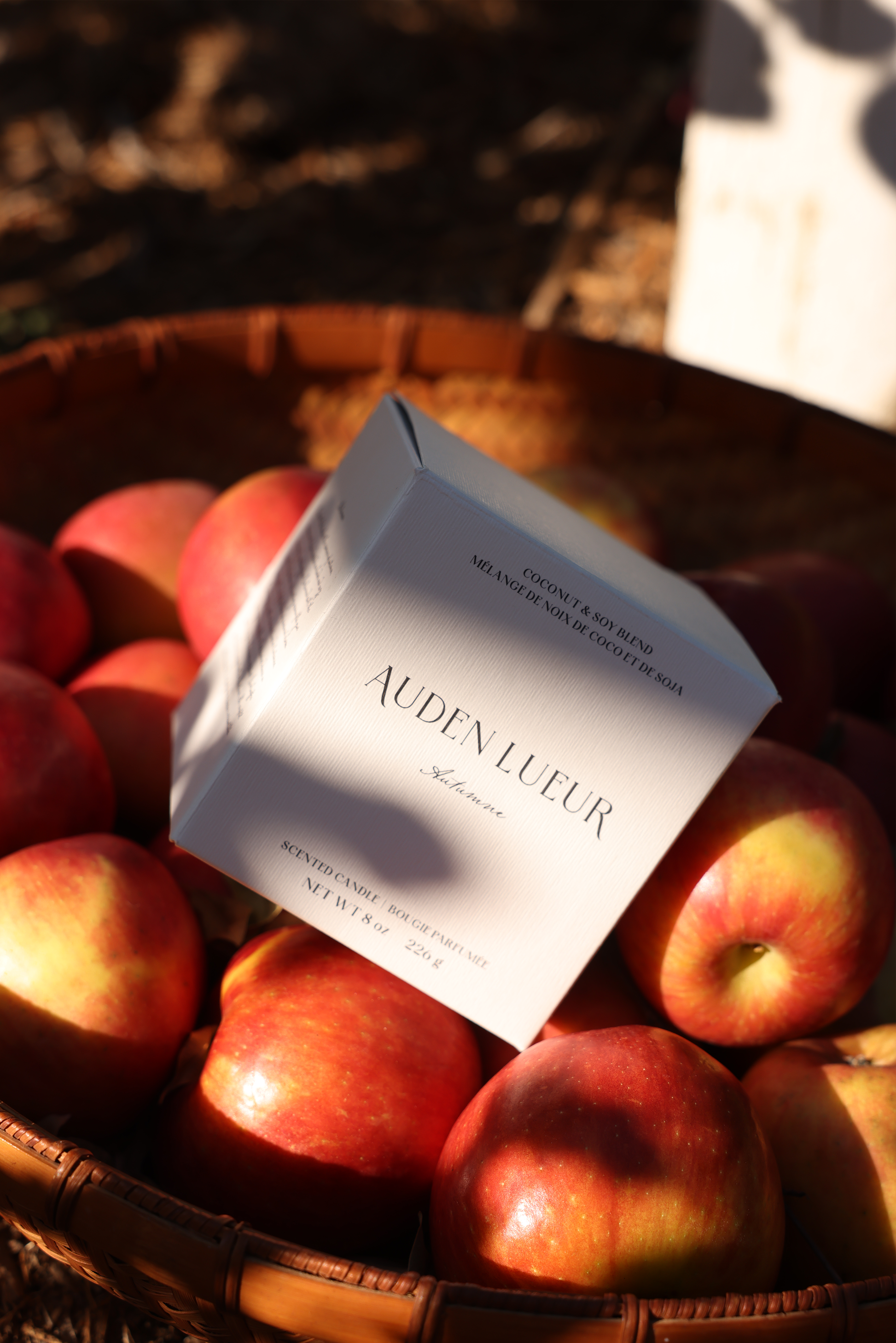 The Autumne candle box leans against apples in a basket.