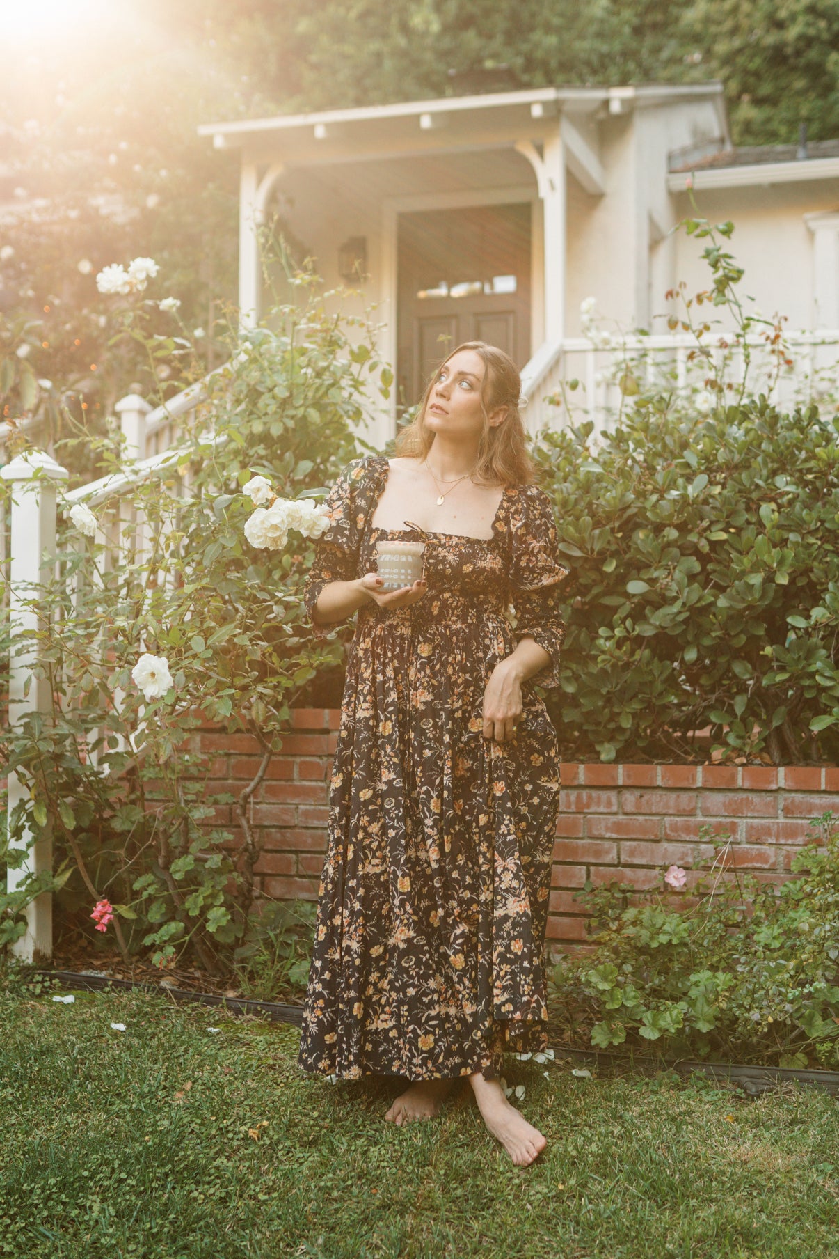 MaCenna Lee holds the Ellori candle, gazing up at the sky, surrounded by the beauty of a garden.