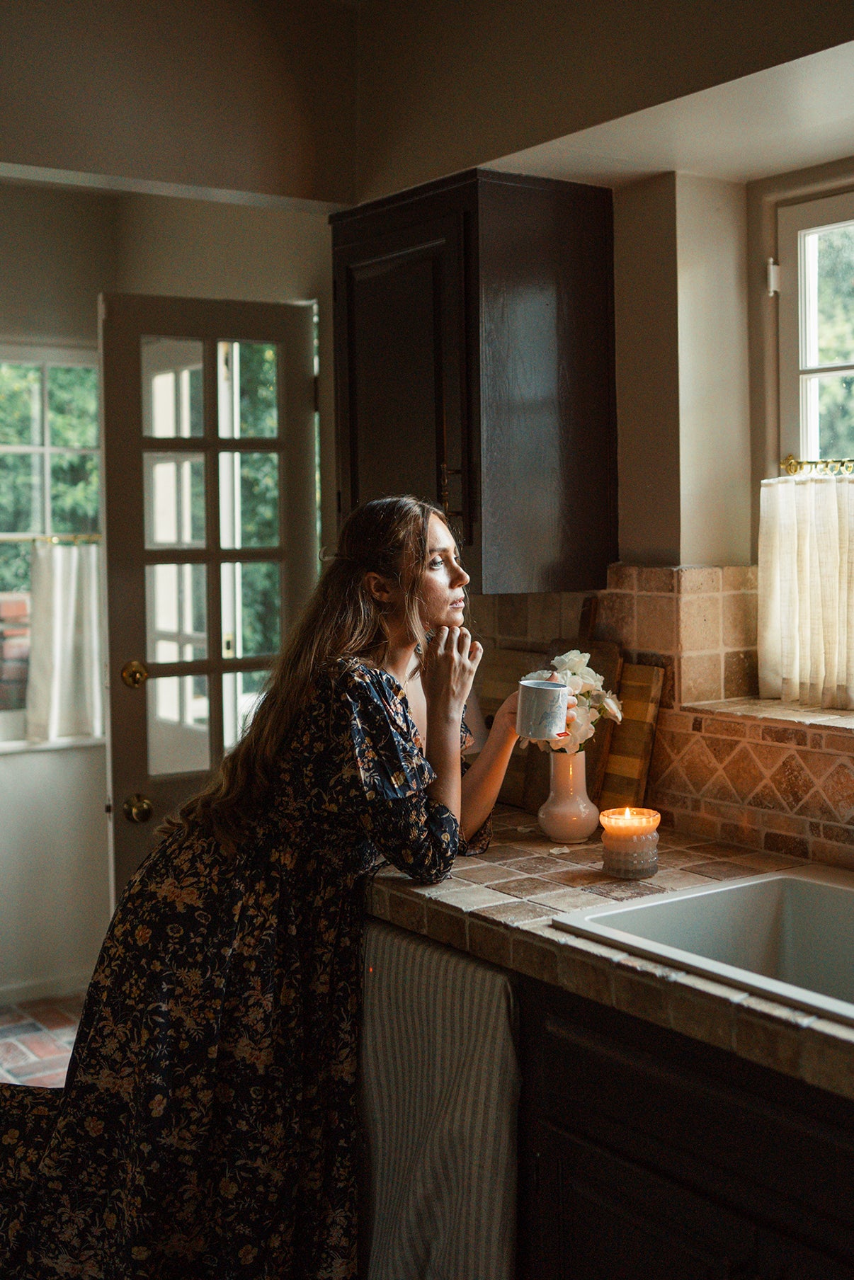 MaCenna Lee holds a cup of coffee in one hand, gazing out the window with the lit Ellori candle nearby.