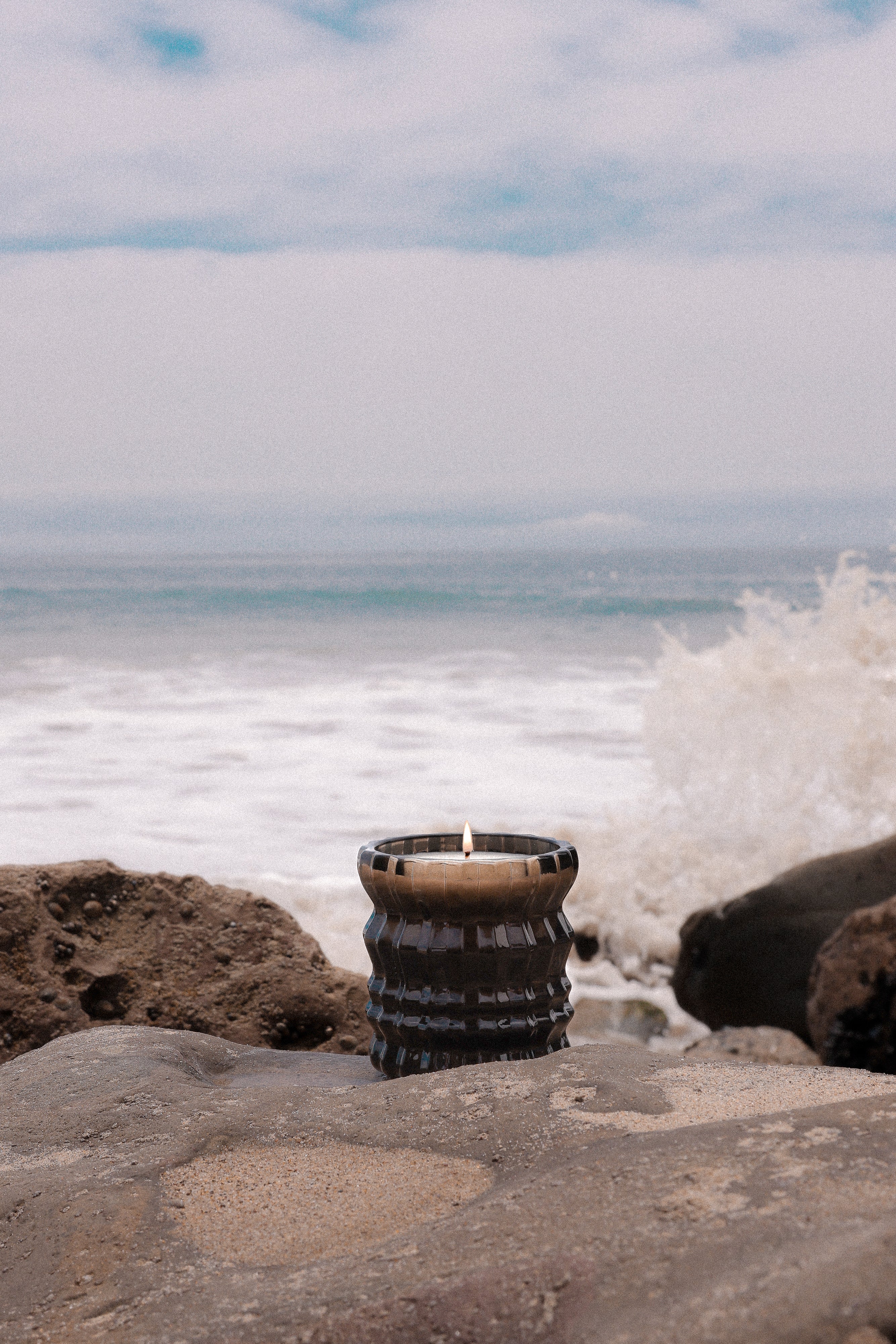The Maris candle sits on a rock before the ocean, its flame flickering as a wave crashes behind it.