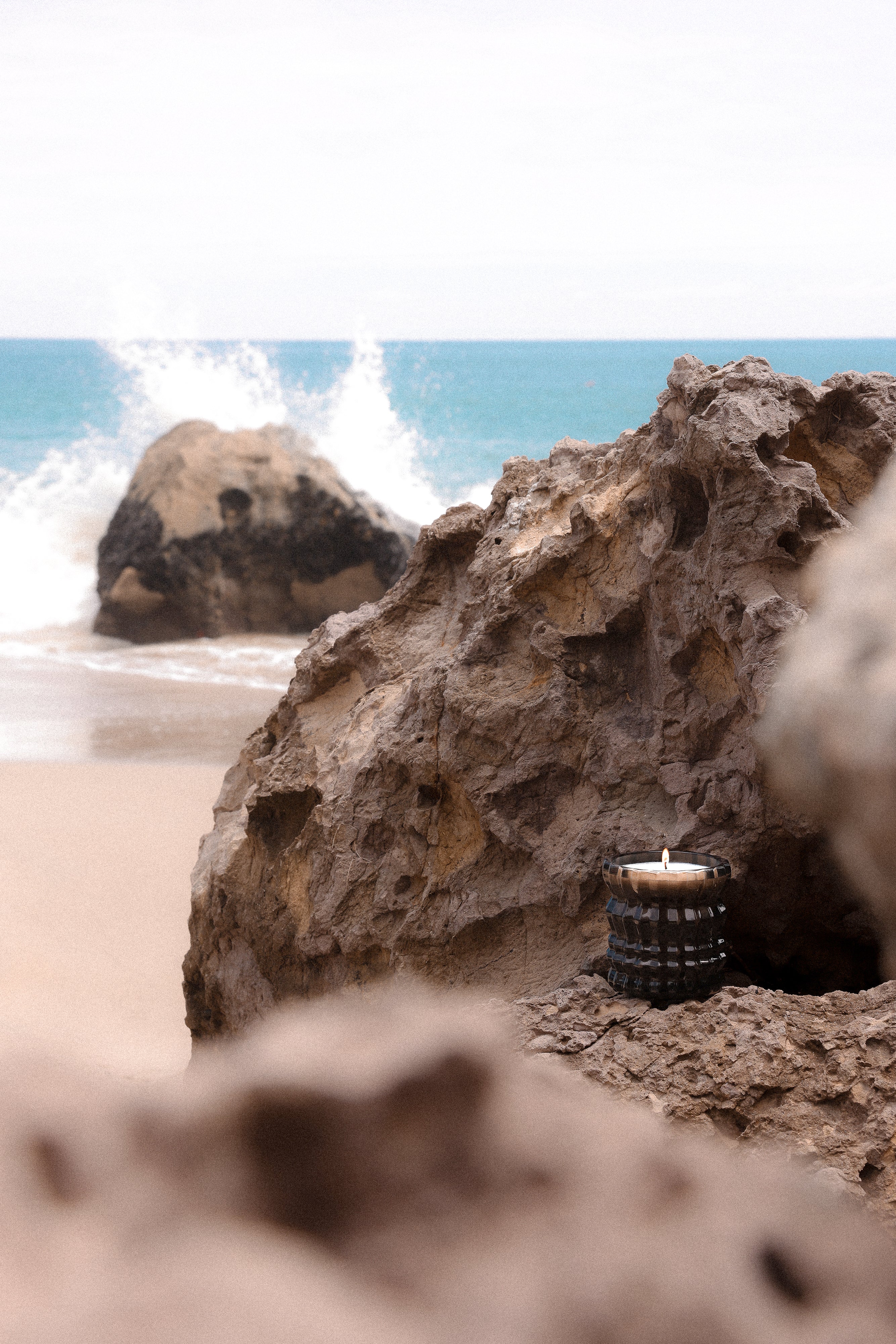 The Maris candle sits on a rock before the ocean, its flame flickering as a wave crashes behind it.